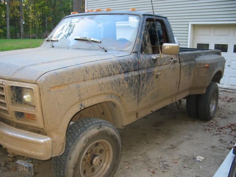 1985 Ford F-350 - This heavily mud-splattered truck is viewed from the front three-quarter angle, showing its dually rear wheels and a portion of the garage…