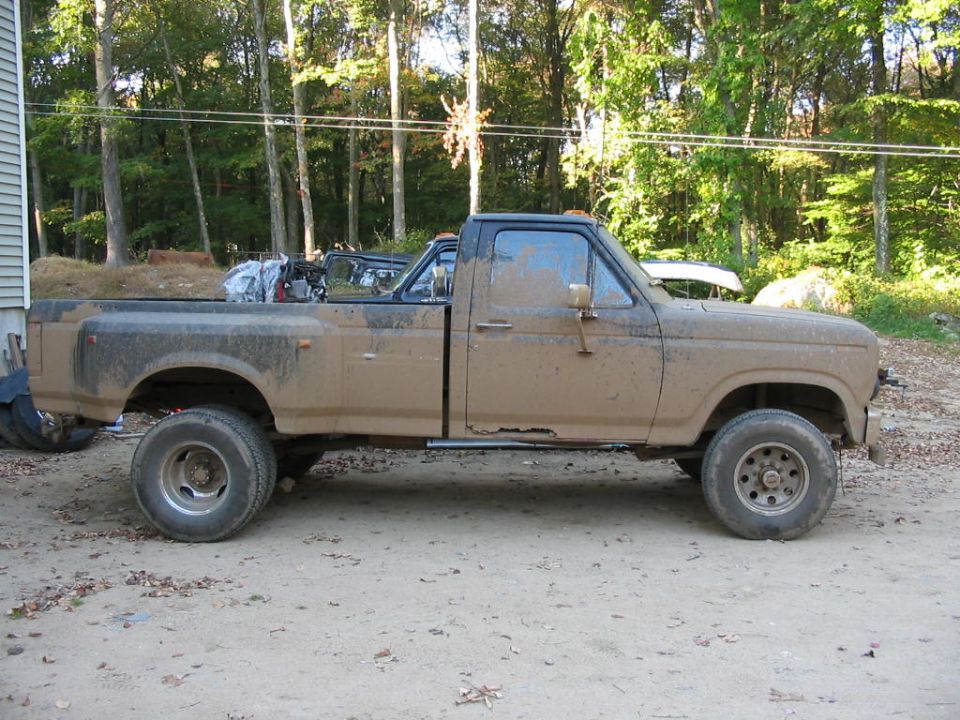 Tan square-body Ford dually 4x4 pickup side profile, mud-splattered, on dirt lot by woods