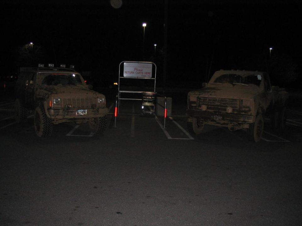 Two muddy trucks parked in a lot at night. The truck on the left appears to be a Jeep Cherokee, and the truck on the right is an older model pickup