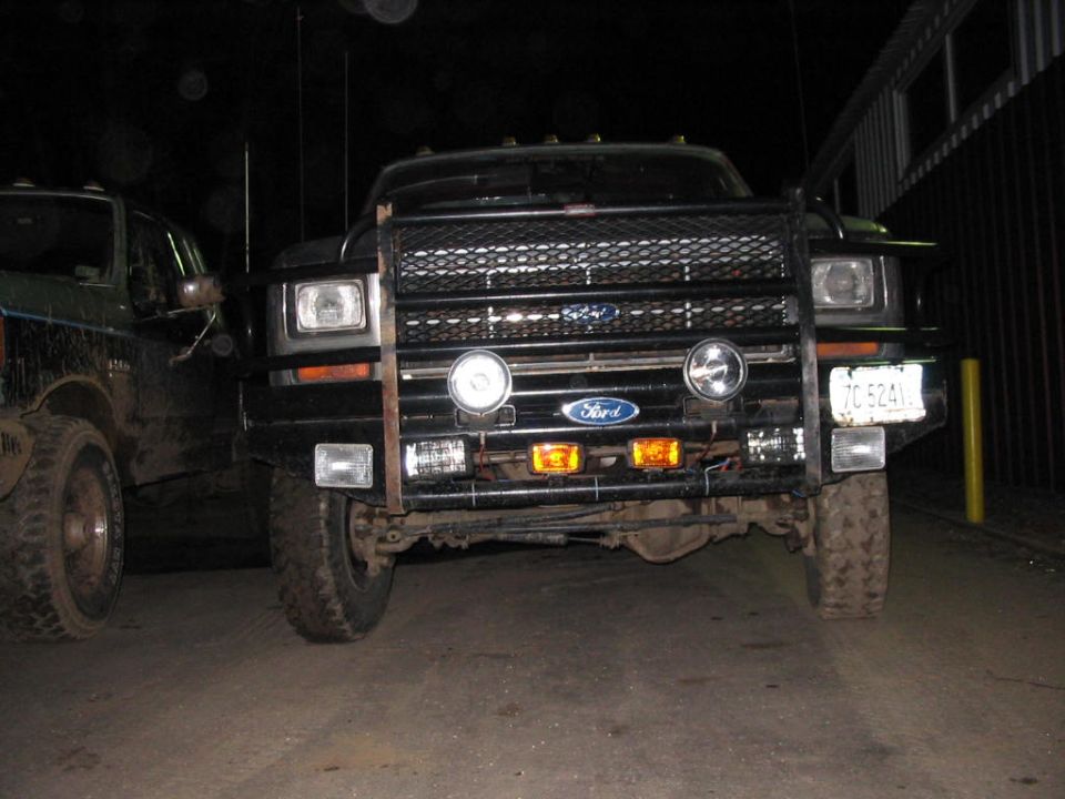 Front view of a black Ford F-Series truck with a brush guard and auxiliary lights. Another truck is partially visible to the left