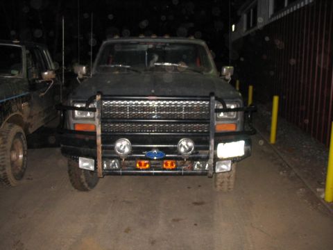 1985 Ford F-350 - A dark Ford pickup truck is parked in front of a wooden fence, with another vehicle to its left.