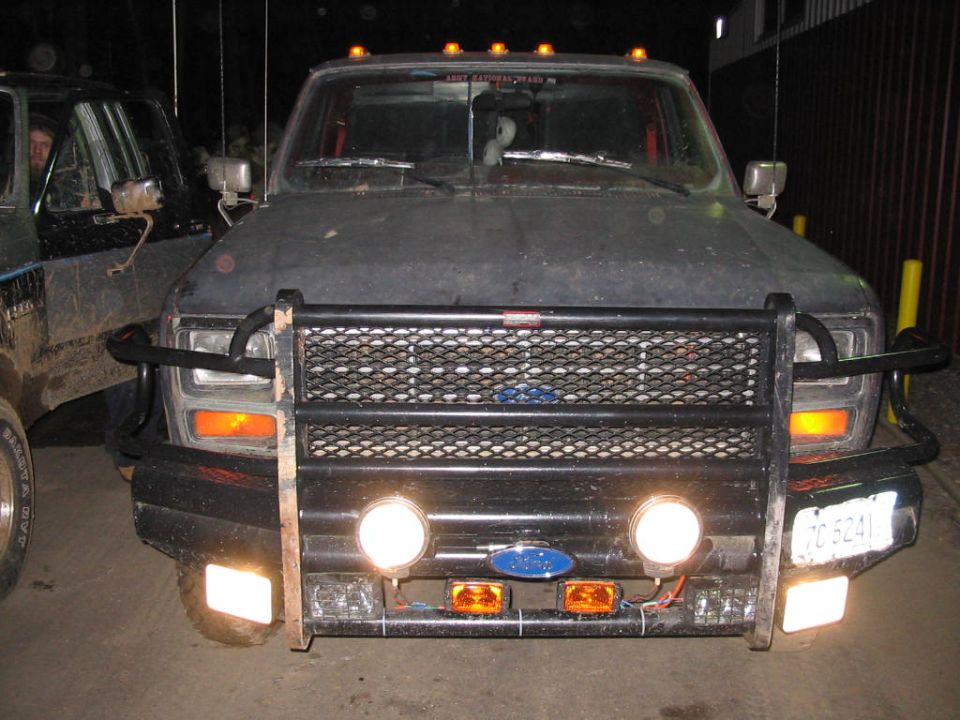 Front view of a dark-colored Ford F-Series truck with a large brush guard and auxiliary lights. Another truck is partially visible to the left. License plate reads TC 5241