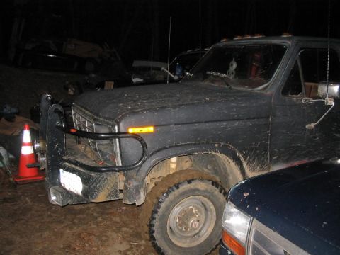 1985 Ford F-350 - A dark, mud-covered pickup truck is parked outdoors at night, with another vehicle partially obscuring the view.