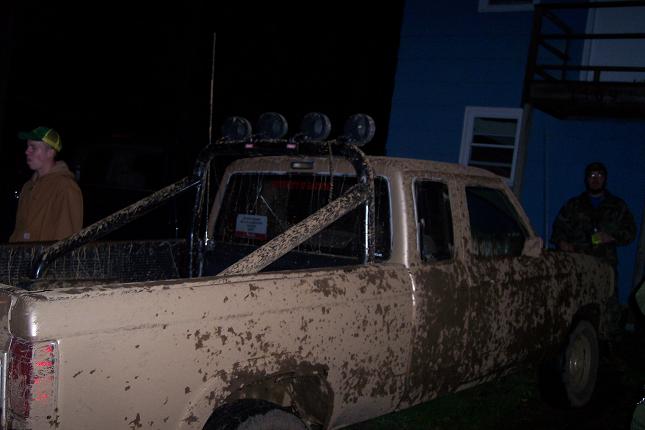 Mud-covered pickup truck with roll bar and lights, parked near a blue building at night
