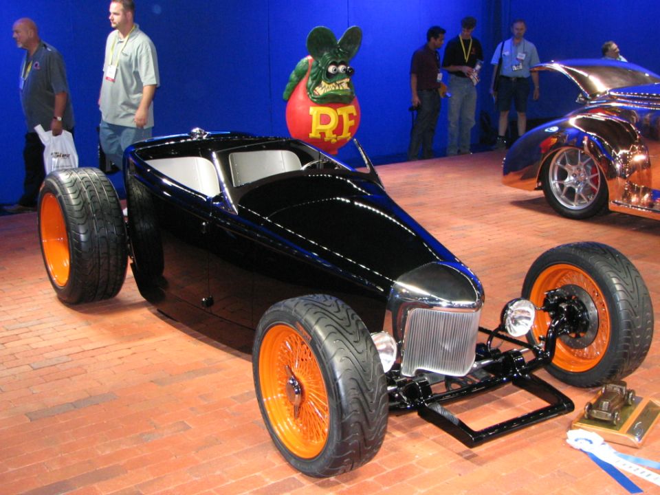 Black hot rod with orange wire wheels and exposed front suspension, seen from a 3/4 front angle