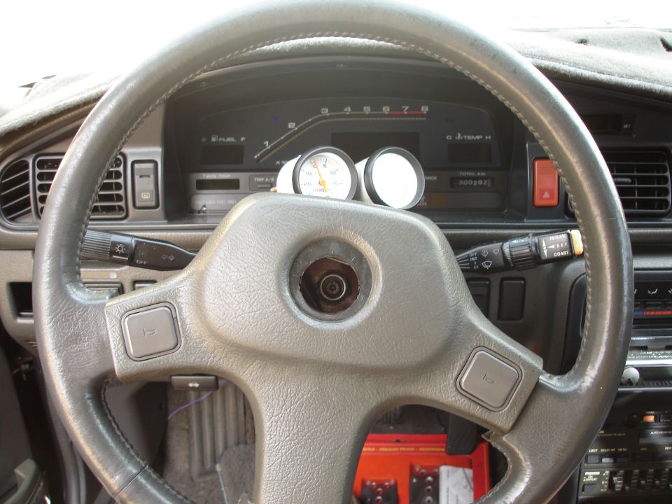 Gray dashboard and steering wheel of a 1989 Mazda MX-6, with aftermarket gauges on the dash