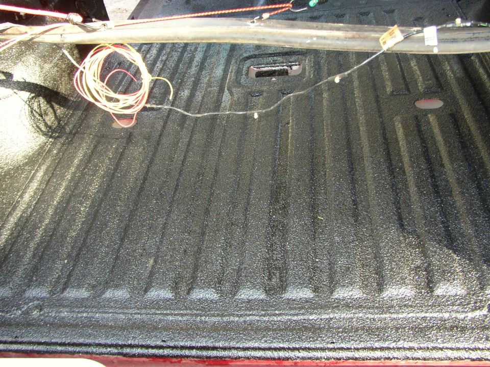 Truck bed interior with black spray-on bedliner and coiled wires. A metal rail runs across the top