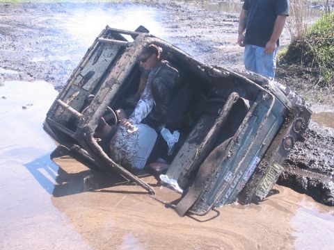 1987 Suzuki Samurai - A vehicle is stuck in the mud, tipped on its side, with a person inside and another person standing nearby.