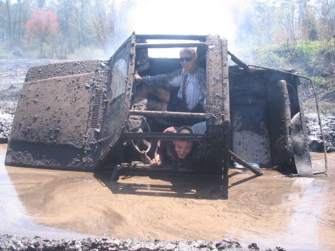 1987 Suzuki Samurai - Two people are inside a heavily mud-covered off-road vehicle that appears to be stuck in a muddy puddle.