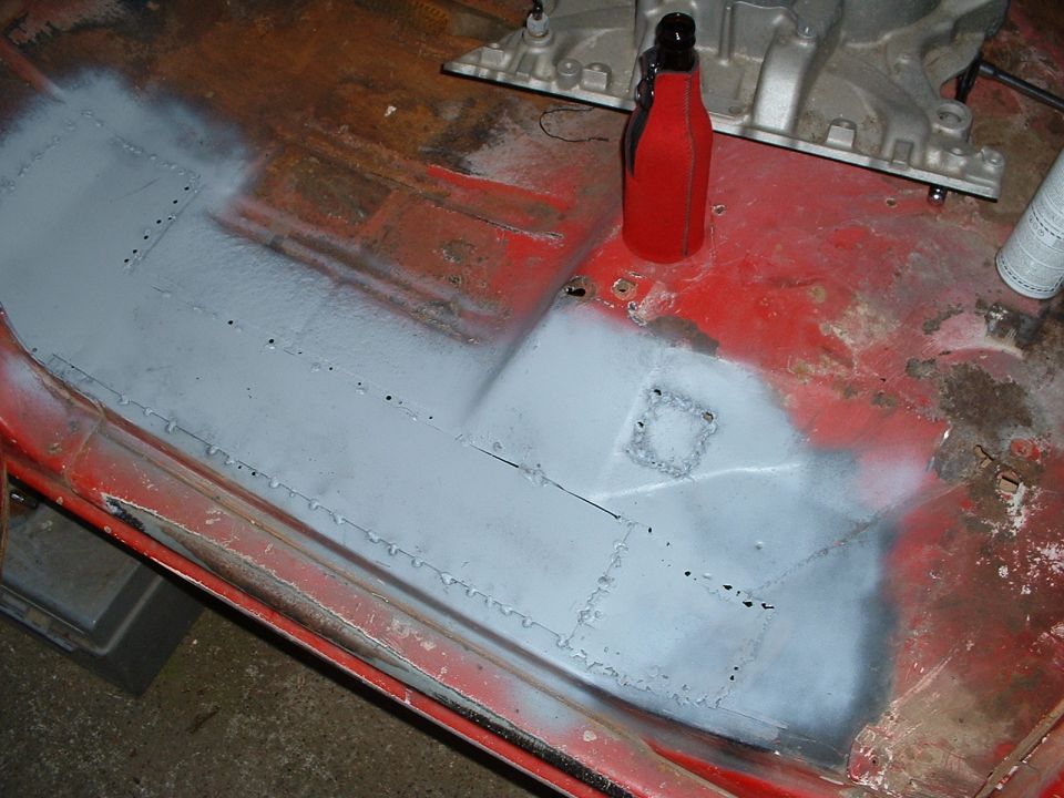 Car floor pan with gray primer applied over patched areas. Red metal visible around edges
