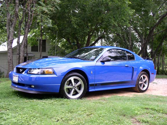 Blue 2000 Ford Mustang parked on grass near a house and trees
