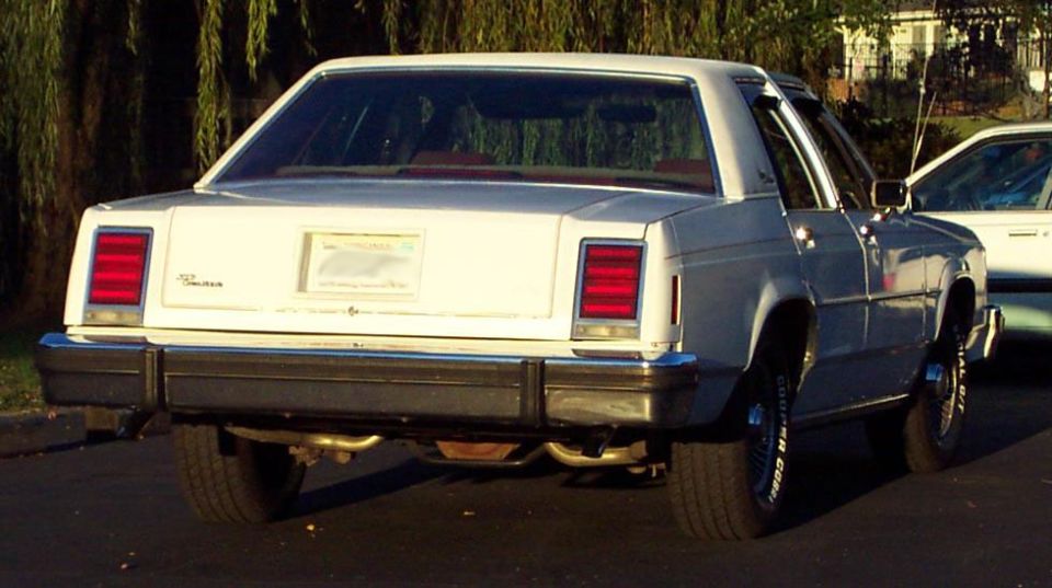 White sedan's rear view, showing rectangular taillights, chrome bumper, and dual exhaust pipes
