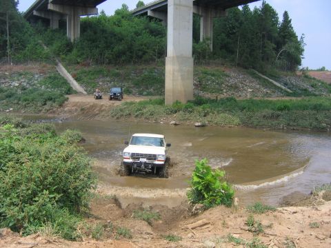 1992 Ford Mustang - A white pickup truck navigates a muddy stream beneath a highway overpass, with other off-road vehicles visible in the distance.
