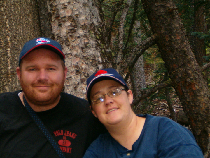 Bearded man and woman in ball caps posing for a close selfie in front of a large pine tree