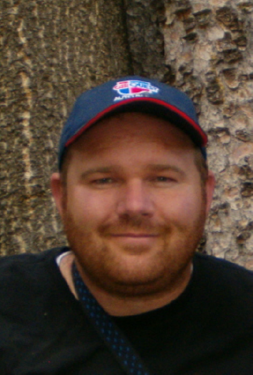 Bearded man in a navy ball cap with red shield logo posing against a rock face