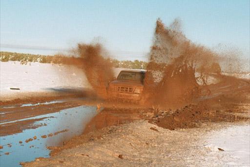Dark Ford truck blasting through a mud hole throwing brown spray on a snowy flat