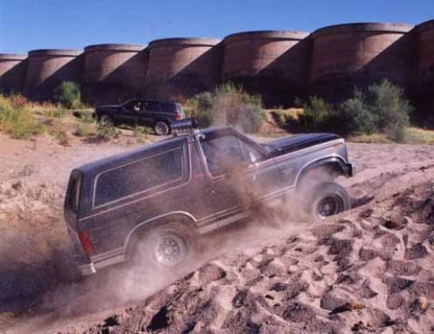 1993 Ford Bronco - A dark gray Ford Bronco navigates a dusty, off-road terrain with a dark SUV in the background.