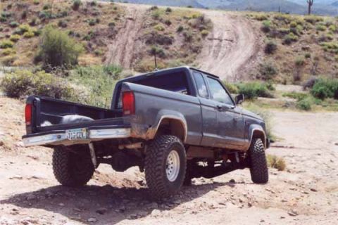 1993 Ford Bronco - A gray pickup truck with large off-road tires is parked on a dusty dirt road with hills in the background.