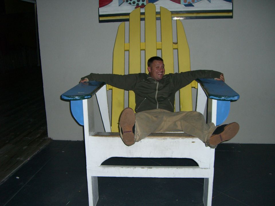 Man with arms outstretched sits in a giant yellow and white Adirondack chair with blue armrests