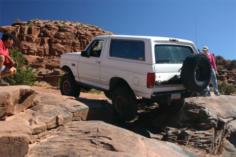1993 Ford Bronco - A white 1993 Ford Bronco navigates a rocky off-road trail under a clear blue sky.