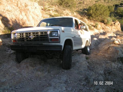 1983 Ford Bronco - White 1983 Ford Bronco navigates a rough, rocky path in a desert landscape.