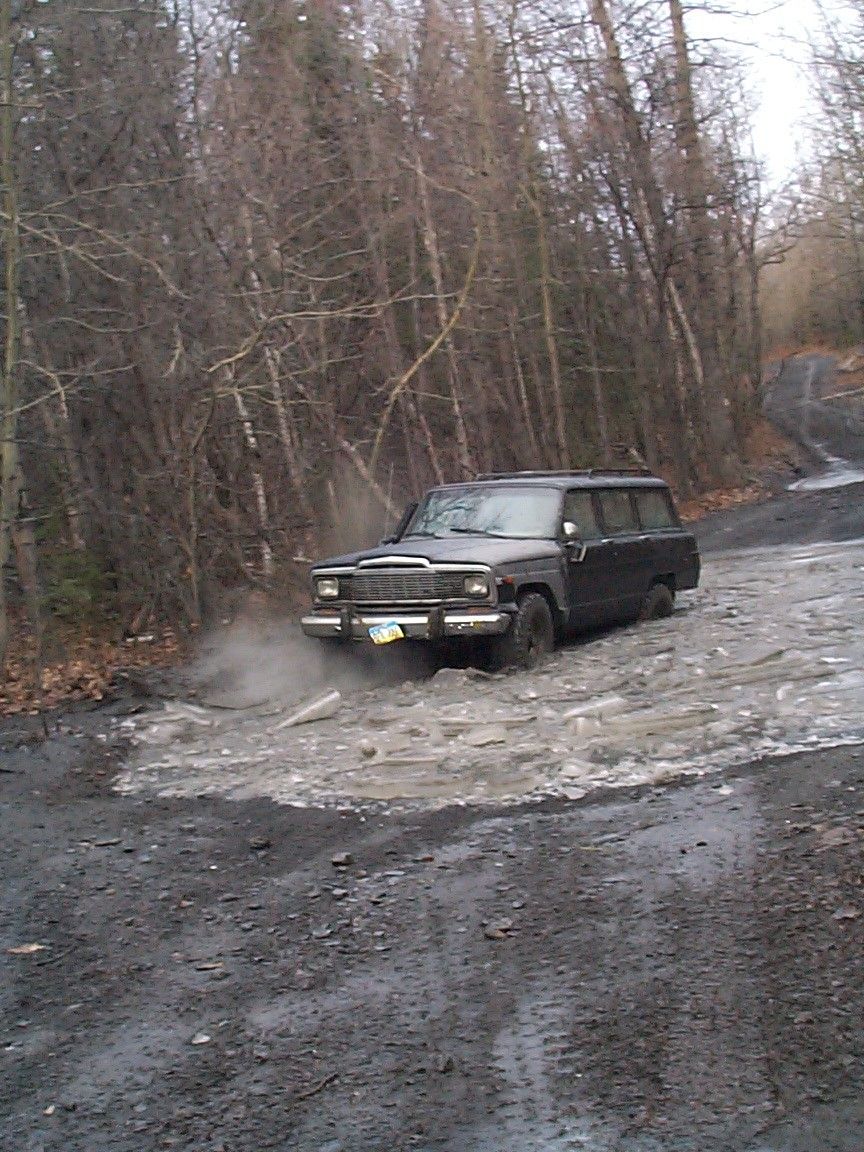 Dark Jeep Wagoneer SUV splashing through mud puddle on a wooded trail
