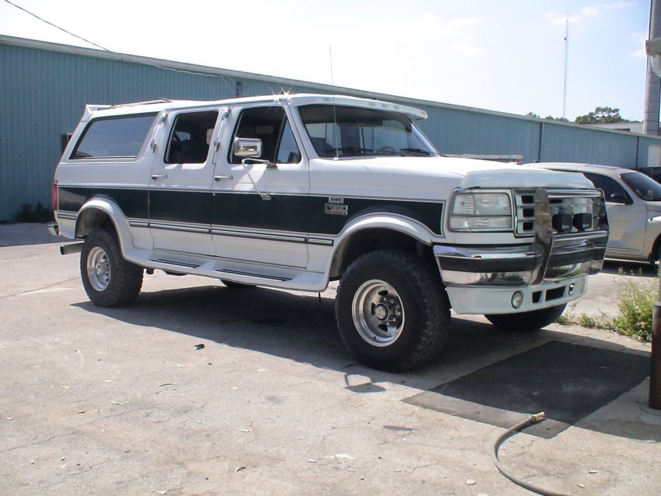 White and black Ford crew-cab conversion SUV with chrome brush guard parked outside a metal building