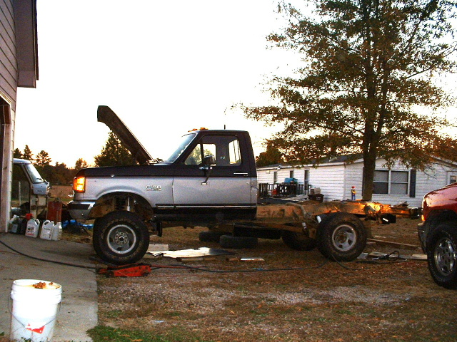 Two-tone Ford F-150 truck chassis with hood open, parked outside a house