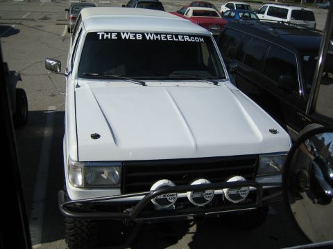 1990 Ford Bronco - white lifted Ford Bronco with off-road lights and a "The Web Wheeler.com" windshield banner, parked front-on in a lot among other vehicles.