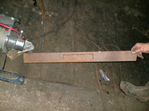 1989 Ford Bronco - A long rusty steel angle-iron bumper face bar lying on a dirt shop floor, with a rectangular slot cut in the center for a fairlead or winch…
