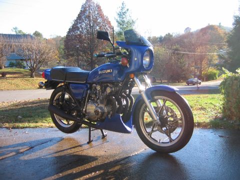 1985 Ford Crown Victoria LTD - A blue Suzuki motorcycle is parked on a wet asphalt surface, with a wooded hillside in the background.