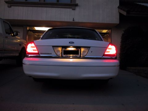 2003 Ford Crown Victoria - The rear of a white 2003 Ford Crown Victoria is visible with its brake lights on, parked in a driveway.