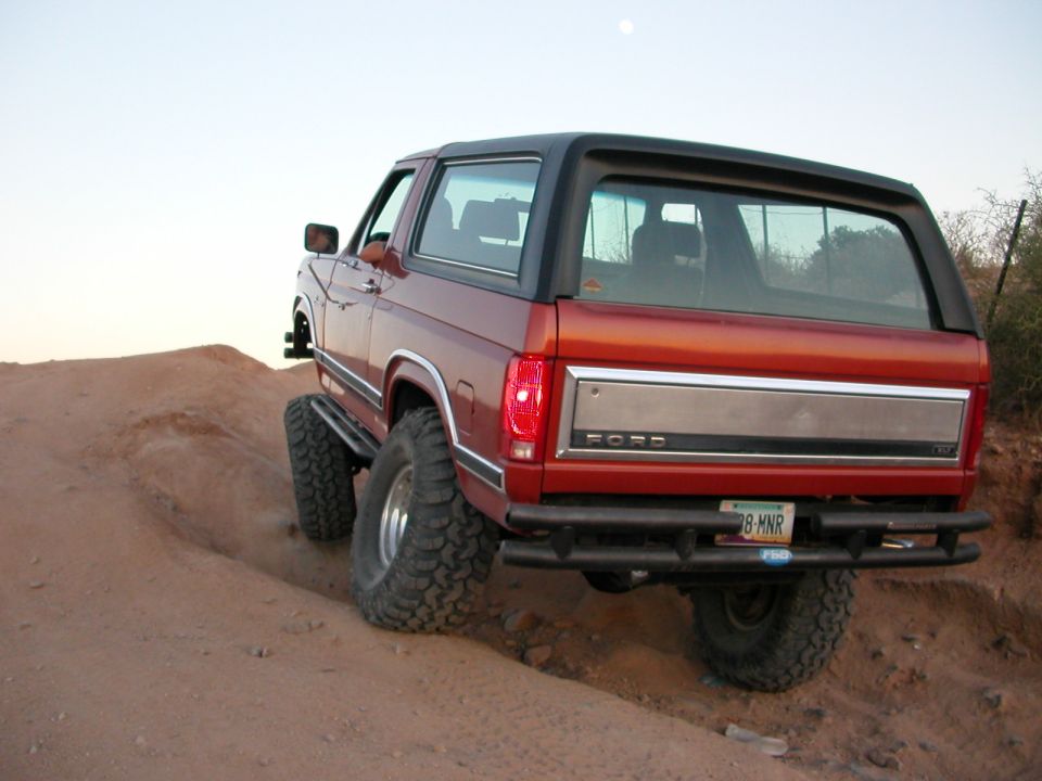 Red Ford Bronco with black top climbing a sandy desert berm, rear view