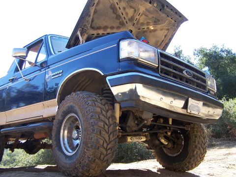 1991 Ford Bronco - Low ground-level shot of a blue and tan two-tone Ford Bronco with the hood propped open, parked on a dirt trail. The lifted front suspensio…