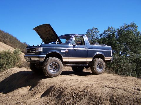 1991 Ford Bronco - Lifted dark blue and tan full-size Ford Bronco with its hardtop removed and hood propped open, posed on a sandy dirt berm against a clear b…