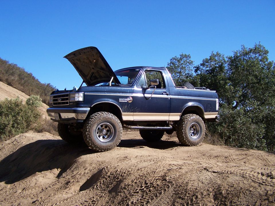 Topless lifted blue Ford Bronco with hood up parked on a dirt hill under blue sky