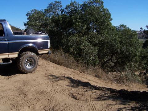 1991 Ford Bronco - A dark blue Ford Bronco with tan trim is shown driving on a dirt hill with trees in the background.