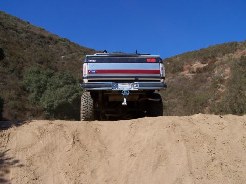 1991 Ford Bronco - This lifted Ford Bronco, seen from the rear, is navigating a sandy incline under a clear blue sky.