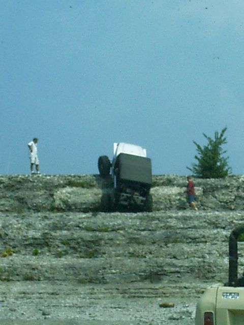 1986 Ford Bronco - An off-road vehicle has tipped onto its side on a steep, rocky incline, with two onlookers observing the scene.