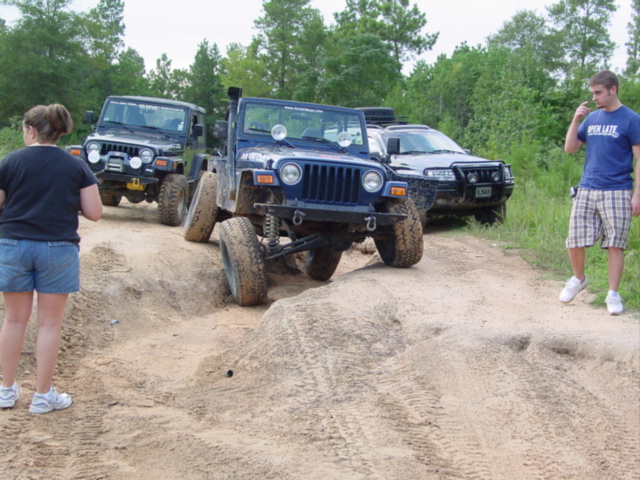 Blue topless Jeep Wrangler flexing suspension over a sand mound on a dirt trail