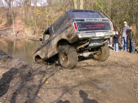 Mud-covered black Ford Bronco attempting to drive through a muddy ditch, rear view