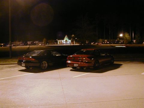 1994 Ford Probe - Two coupes are parked in a lot at night, with buildings and lights in the background.