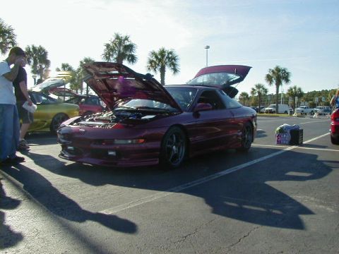 1994 Ford Probe - A group of people stand near a maroon Ford Probe with its hood and trunk open, parked in a lot with other vehicles.