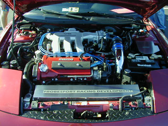 Engine bay of a red car with a V6 engine, blue spark plug wires, and a "PROBESPORT RACING DEVELOPMENT" plate