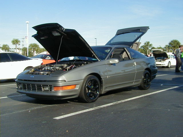 Gray coupe with hood and trunk open, parked in a lot with other cars. Engine is visible