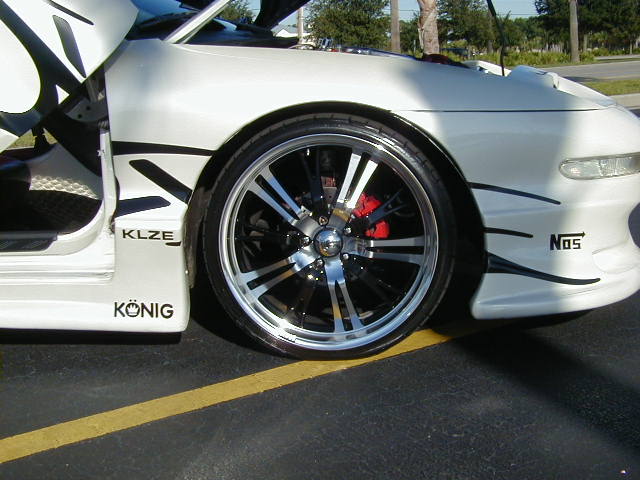 White 1994 Ford Probe with custom chrome wheels, red brake calipers, and black racing stripes. The driver's side door is open