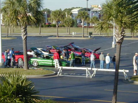1994 Ford Probe - A group of people gather around a green 1994 Ford Probe with white racing stripes parked in a lot with other vehicles.