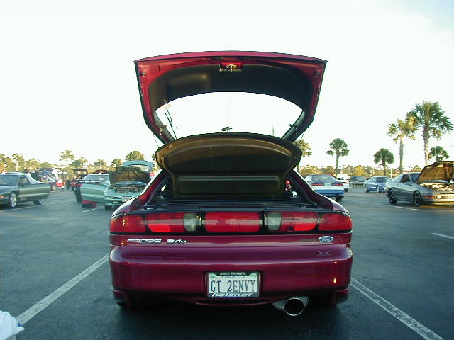 Red hatchback with trunk and hood open, parked at an outdoor car meet. Other cars visible in background