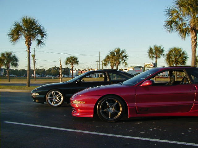 Red and black Ford Probes parked on asphalt with palm trees in background. Red car has GT badge