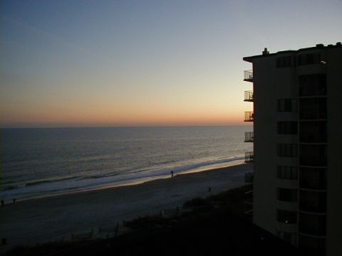 1994 Ford Probe - A tranquil beach scene unfolds with gentle waves lapping the shore under a colorful sunset, beside a multi-story building.